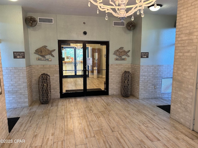 interior space with an inviting chandelier, brick wall, light wood-type flooring, a textured ceiling, and french doors