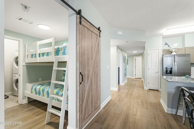 bedroom with stainless steel refrigerator with ice dispenser, a barn door, light hardwood / wood-style flooring, and stacked washer and clothes dryer