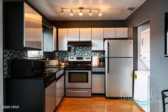 kitchen with stacked washer and clothes dryer, a textured ceiling, appliances with stainless steel finishes, light hardwood / wood-style floors, and extractor fan