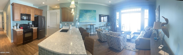 kitchen with a sink, brown cabinets, black appliances, dark wood finished floors, and a center island with sink