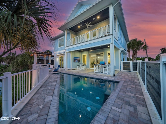 back house at dusk featuring a pergola, a balcony, ceiling fan, a patio, and a fenced in pool