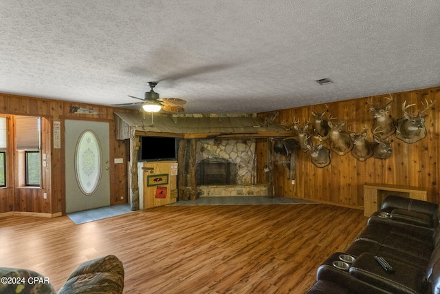 living room featuring wood walls, hardwood / wood-style floors, a stone fireplace, ceiling fan, and a textured ceiling