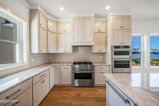 kitchen with tasteful backsplash, light stone countertops, appliances with stainless steel finishes, light wood-type flooring, and cream cabinets