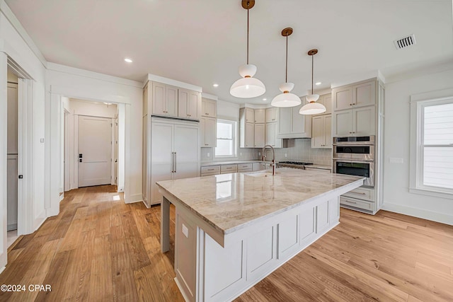 kitchen with tasteful backsplash, hanging light fixtures, an island with sink, light stone countertops, and light hardwood / wood-style floors