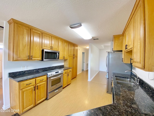 kitchen with sink, stainless steel appliances, dark stone counters, a textured ceiling, and light wood-type flooring