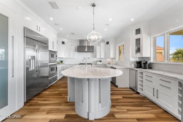 kitchen featuring hardwood / wood-style floors, an island with sink, backsplash, wall chimney range hood, and appliances with stainless steel finishes