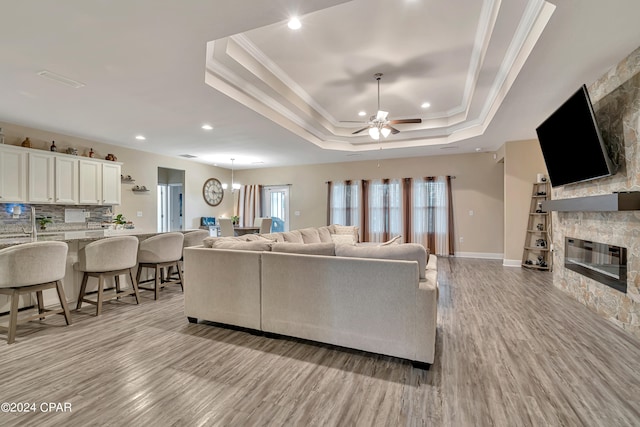 living room with light hardwood / wood-style floors, ceiling fan with notable chandelier, a tray ceiling, a stone fireplace, and ornamental molding