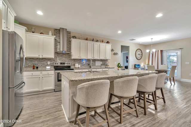 kitchen with light hardwood / wood-style floors, appliances with stainless steel finishes, an island with sink, wall chimney exhaust hood, and backsplash