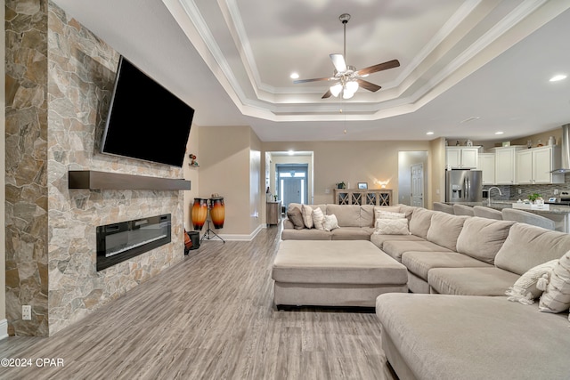 living room featuring crown molding, light hardwood / wood-style floors, a stone fireplace, ceiling fan, and a raised ceiling