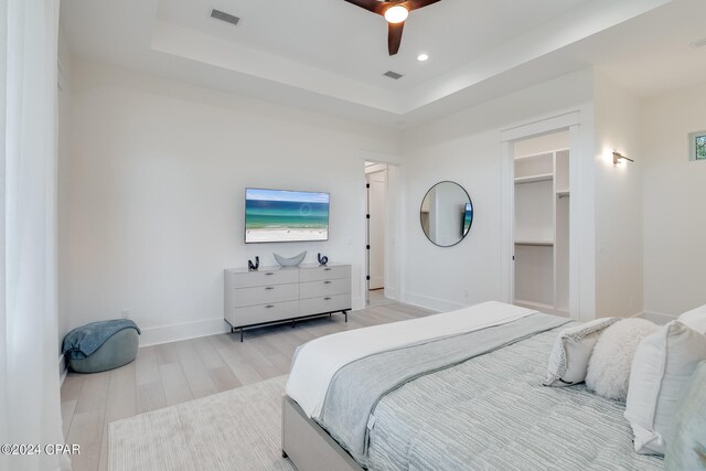 bedroom with light wood-type flooring, a raised ceiling, a walk in closet, and ceiling fan