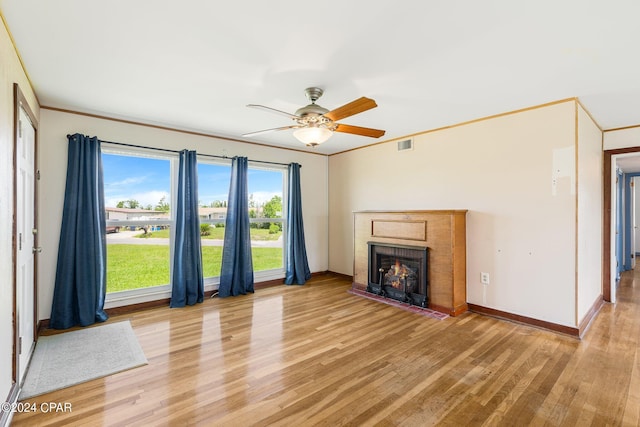 unfurnished living room featuring a warm lit fireplace, baseboards, ceiling fan, ornamental molding, and light wood-type flooring