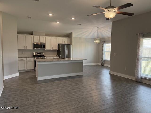 kitchen with dark wood-type flooring, white cabinets, ceiling fan with notable chandelier, a center island with sink, and appliances with stainless steel finishes