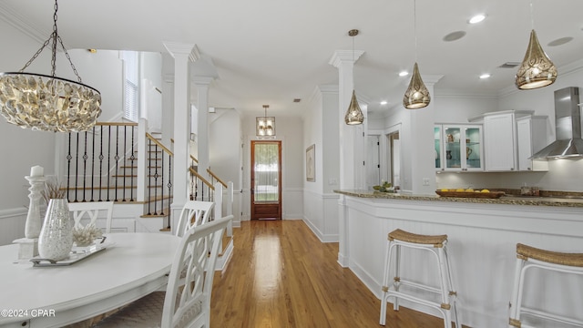 kitchen with wall chimney range hood, light hardwood / wood-style floors, white cabinetry, hanging light fixtures, and ornate columns