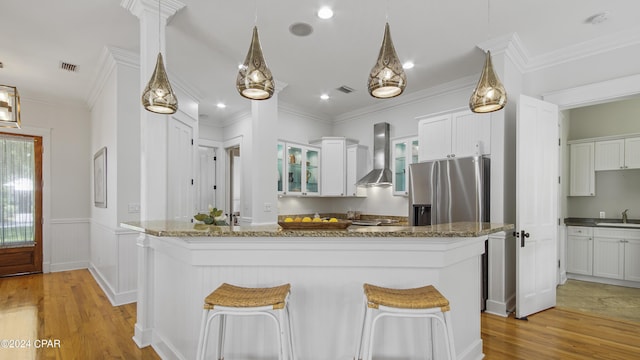 kitchen with stainless steel fridge, hanging light fixtures, wall chimney range hood, white cabinets, and a center island