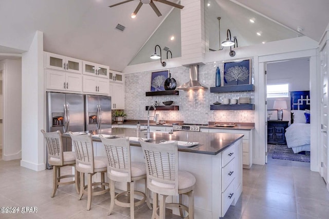 kitchen with stainless steel refrigerator with ice dispenser, white cabinetry, a center island with sink, wall chimney range hood, and backsplash