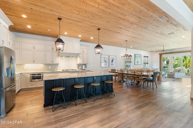 kitchen with light wood-type flooring, a center island with sink, appliances with stainless steel finishes, decorative light fixtures, and tasteful backsplash