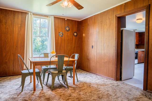 dining space with wood walls, light colored carpet, ornamental molding, and ceiling fan