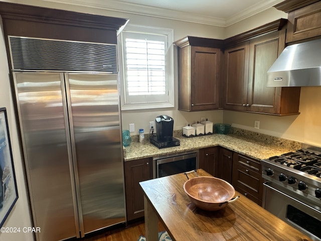 kitchen with dark brown cabinets, high end appliances, extractor fan, light stone counters, and ornamental molding