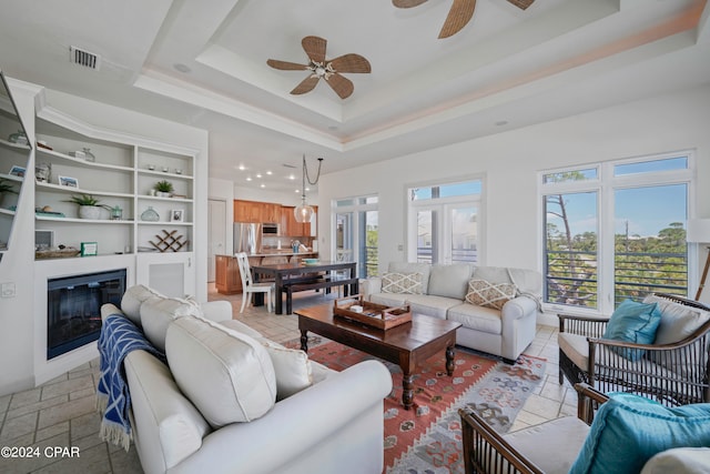 living room featuring built in shelves, a raised ceiling, plenty of natural light, and ceiling fan