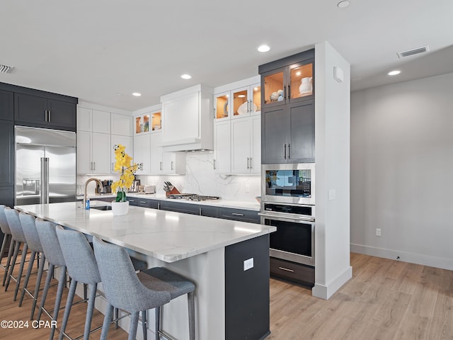 kitchen with light stone counters, built in appliances, a kitchen island with sink, and light hardwood / wood-style flooring