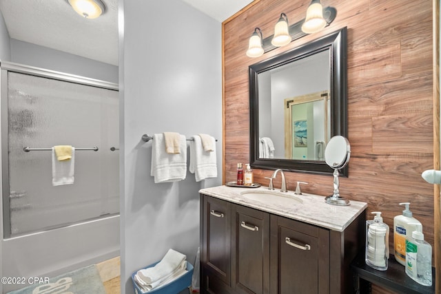 bathroom with a textured ceiling, wooden walls, enclosed tub / shower combo, and vanity