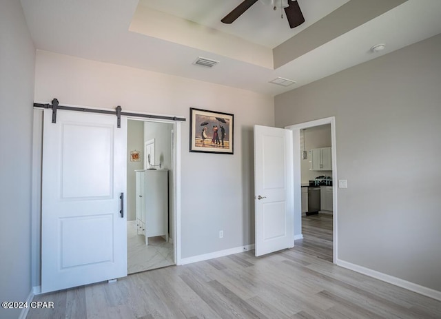 unfurnished bedroom featuring light hardwood / wood-style flooring, ceiling fan, a barn door, connected bathroom, and a tray ceiling