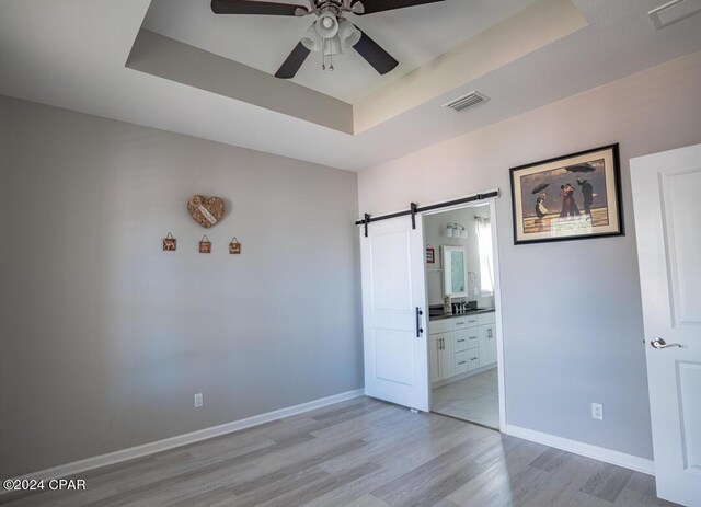 unfurnished bedroom with ceiling fan, a barn door, a tray ceiling, and light hardwood / wood-style floors