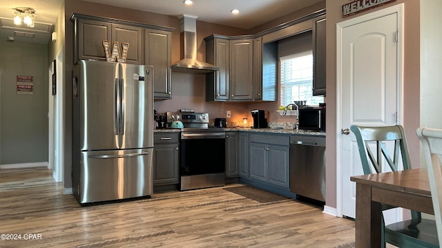 kitchen featuring light hardwood / wood-style flooring, appliances with stainless steel finishes, wall chimney range hood, light stone countertops, and gray cabinetry