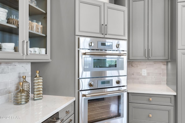 kitchen with gray cabinetry, decorative backsplash, light stone counters, and stainless steel double oven