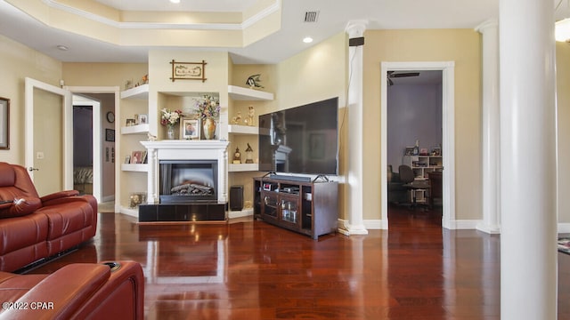 living room featuring ornamental molding, built in shelves, dark wood-type flooring, a raised ceiling, and decorative columns