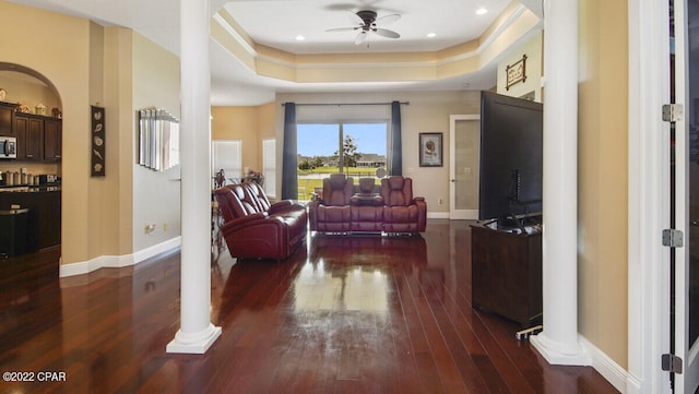 living room with ceiling fan, a tray ceiling, dark wood-type flooring, and decorative columns