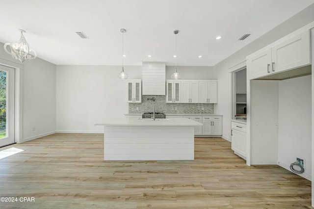 kitchen with wall chimney range hood, decorative light fixtures, a kitchen island with sink, and white cabinets