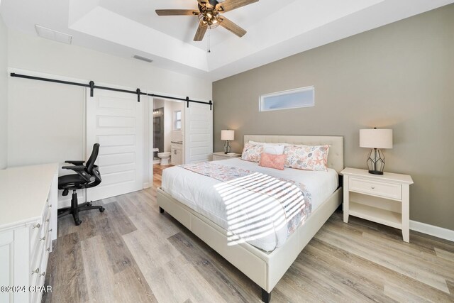 bedroom featuring connected bathroom, light hardwood / wood-style flooring, a tray ceiling, and a barn door