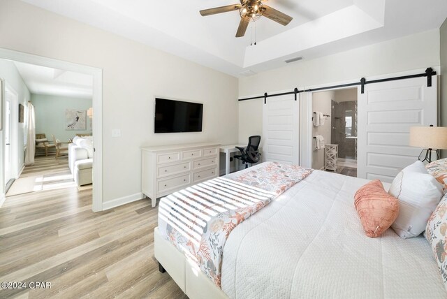 bedroom featuring light hardwood / wood-style floors, a raised ceiling, a barn door, ceiling fan, and ensuite bathroom