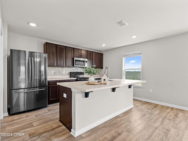 kitchen featuring a kitchen breakfast bar, a kitchen island with sink, dark brown cabinetry, stainless steel appliances, and light hardwood / wood-style flooring