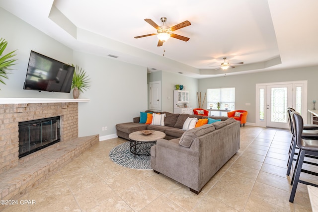 living area featuring light tile patterned floors, baseboards, a ceiling fan, a tray ceiling, and a fireplace