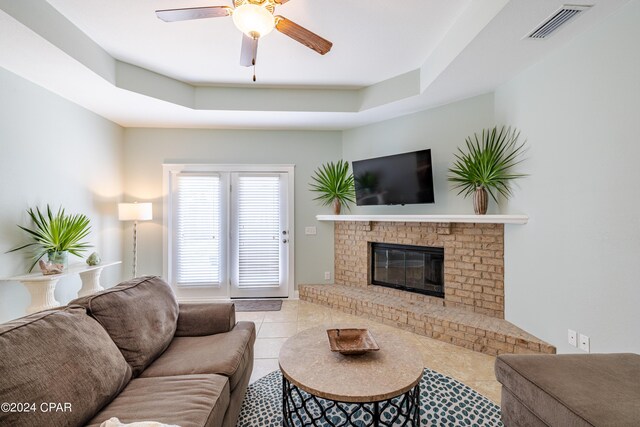 living room featuring a raised ceiling, a brick fireplace, light tile patterned floors, and ceiling fan