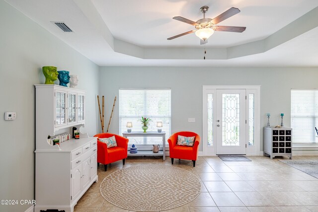 living room featuring ceiling fan, a tray ceiling, a brick fireplace, and light tile patterned floors