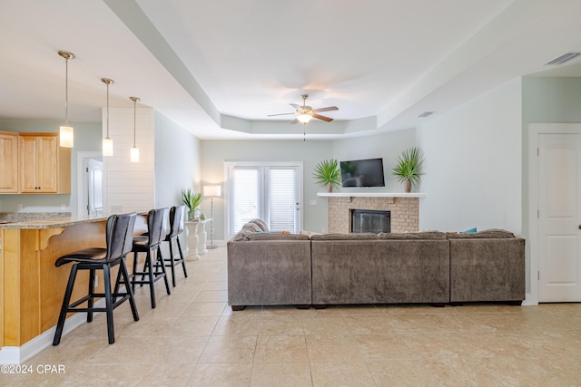 tiled living room with ceiling fan, a raised ceiling, and a brick fireplace