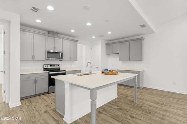 kitchen with gray cabinets, stainless steel appliances, an island with sink, sink, and light hardwood / wood-style flooring