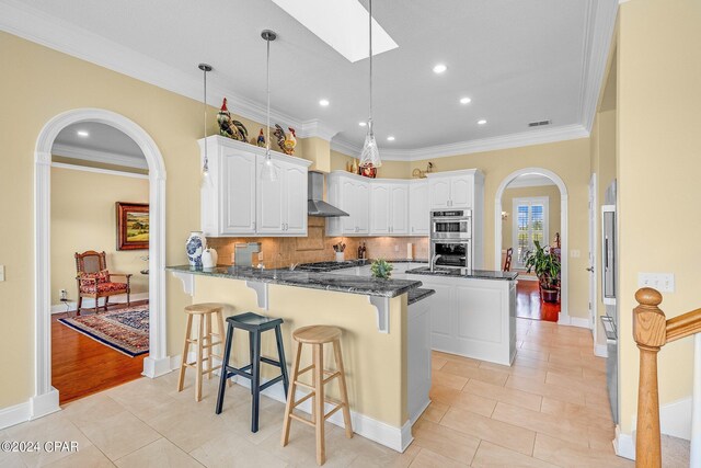 kitchen featuring white cabinetry, a skylight, kitchen peninsula, appliances with stainless steel finishes, and wall chimney range hood