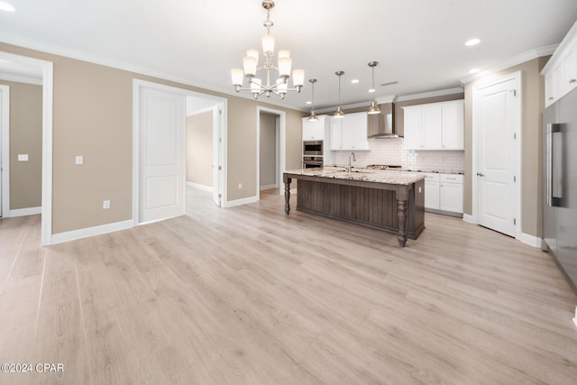 kitchen with hanging light fixtures, light hardwood / wood-style floors, white cabinetry, wall chimney exhaust hood, and a kitchen island with sink