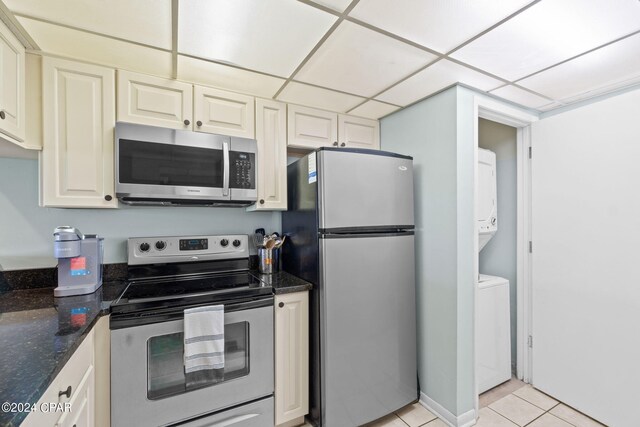 kitchen with stacked washer and clothes dryer, dark stone countertops, light tile patterned floors, appliances with stainless steel finishes, and a paneled ceiling