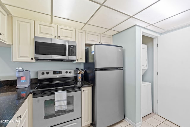 kitchen with appliances with stainless steel finishes, light tile patterned flooring, a paneled ceiling, stacked washer and dryer, and dark stone countertops