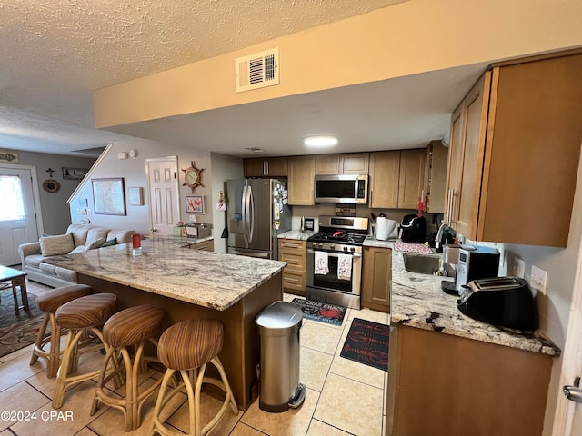 kitchen with a textured ceiling, stainless steel appliances, kitchen peninsula, light stone countertops, and a breakfast bar