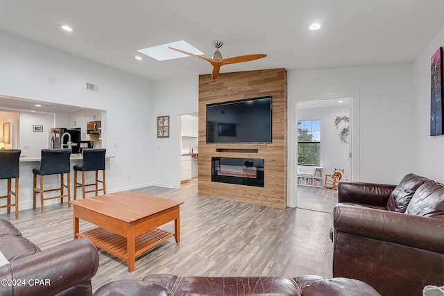 living room with a large fireplace, ceiling fan, vaulted ceiling with skylight, and light hardwood / wood-style floors