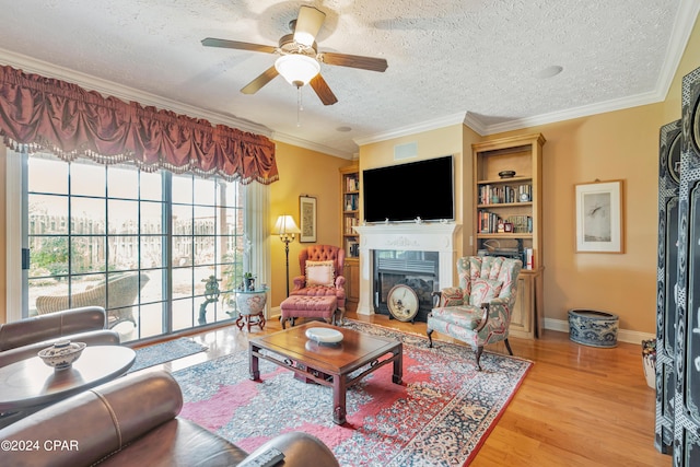 living room featuring light wood-type flooring, a ceiling fan, a textured ceiling, a glass covered fireplace, and crown molding