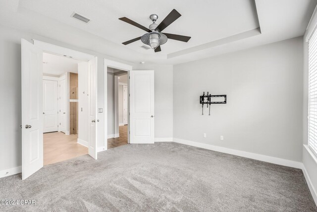 unfurnished bedroom with ceiling fan, light colored carpet, a raised ceiling, and multiple windows