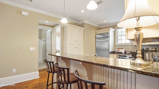 kitchen with light hardwood / wood-style floors, hanging light fixtures, built in fridge, dark stone countertops, and a kitchen bar