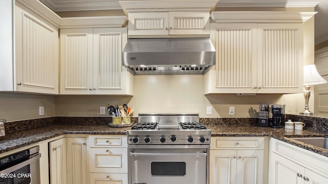 kitchen with ornamental molding, cream cabinets, appliances with stainless steel finishes, range hood, and dark stone counters